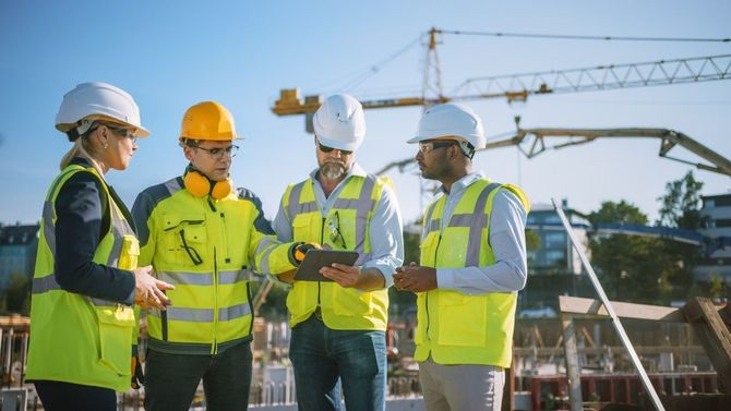 Four construction workers in safety vests and hard hats discussing at a building site under a crane.