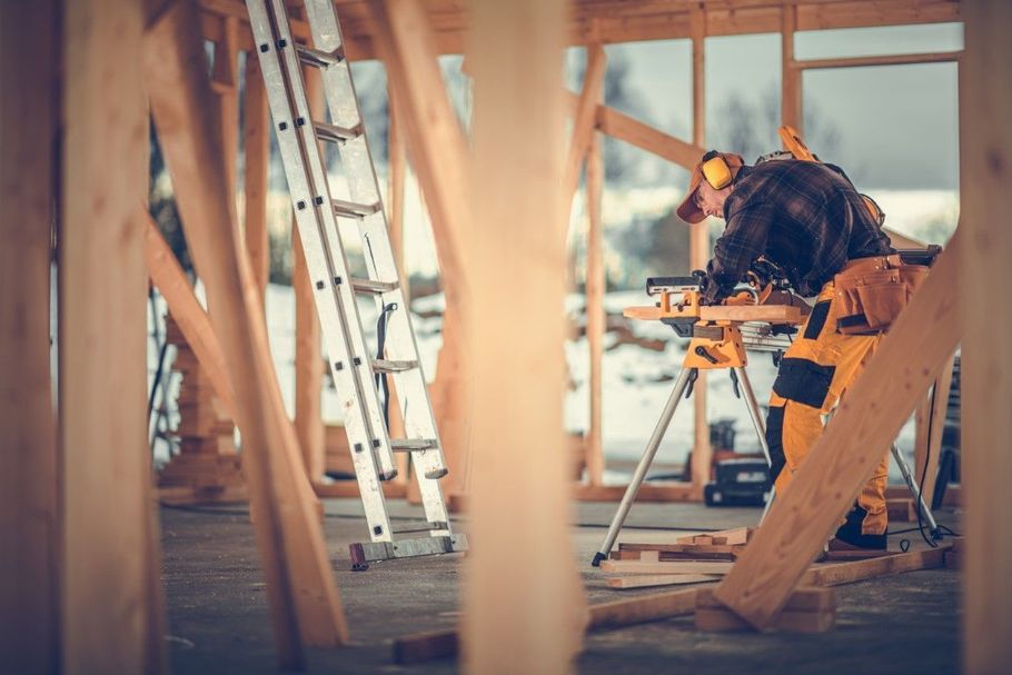 Worker using a miter saw in a sunlit construction site, with ladders and wooden framing nearby.