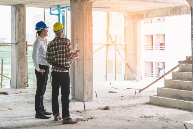 Two workers in hard hats inspecting an unfinished building interior with stairs and scaffolding.