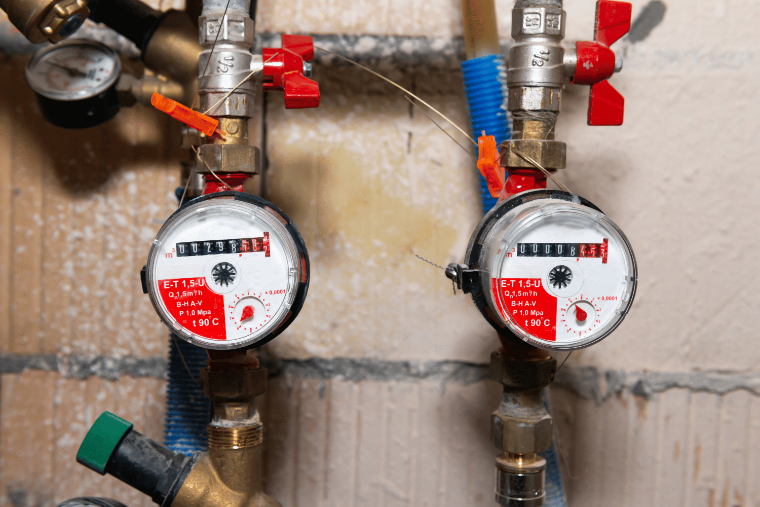 Two water meters with red valves and colored pipes on a utility wall