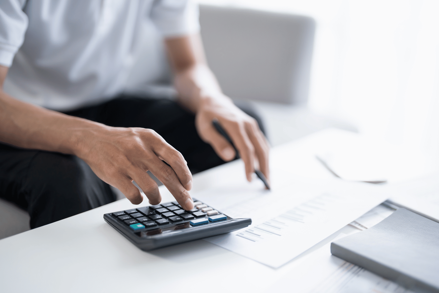 Person using a calculator at a white desk with papers and a laptop