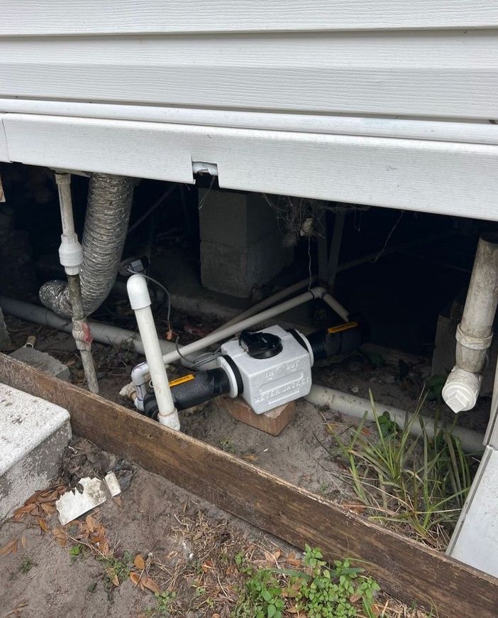 Open crawl space under a house with exposed pipes and a white utility pump.