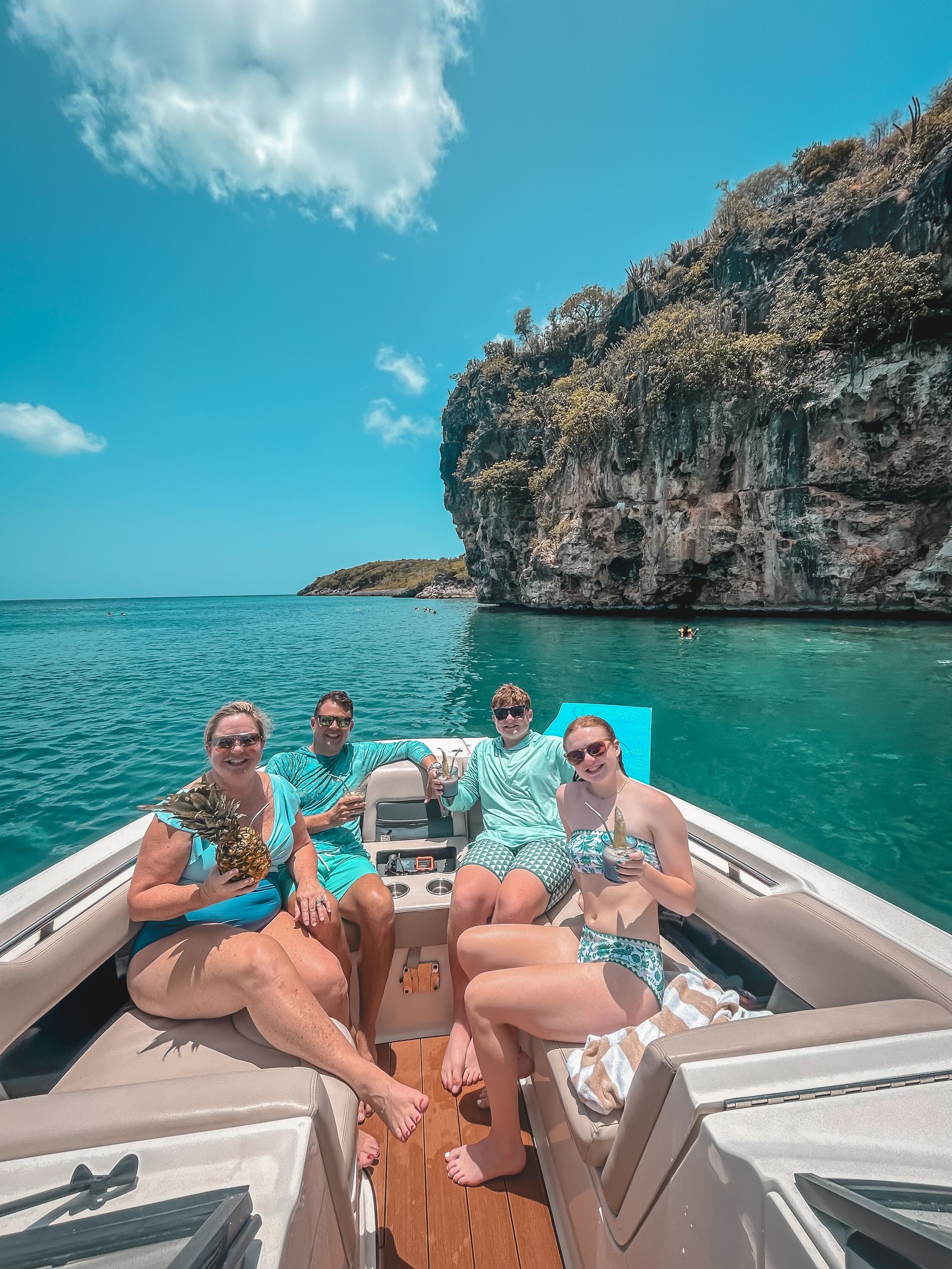 Four people on a boat in turquoise water near a rocky cliff on a sunny day.