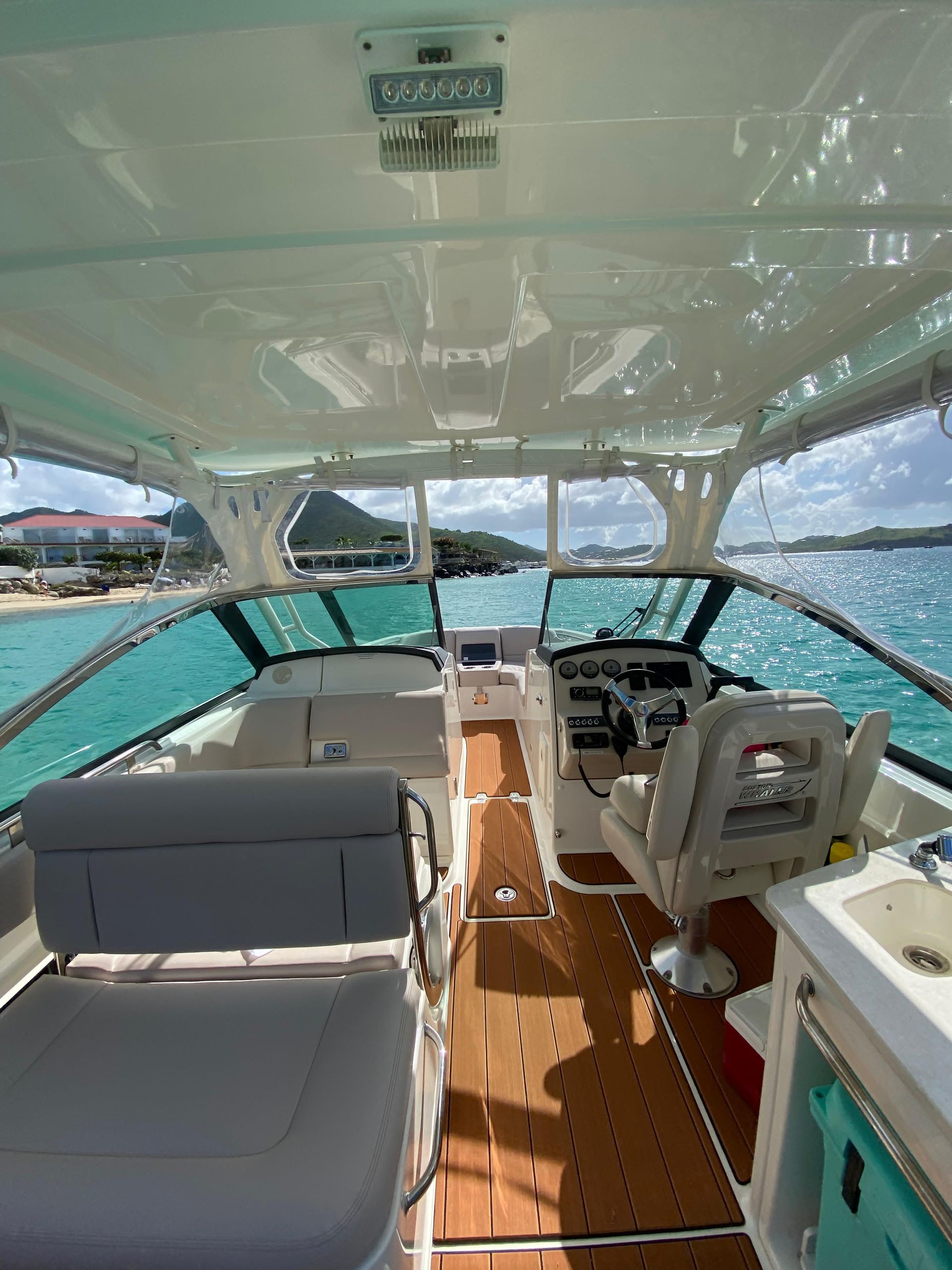 Interior of a boat with wooden floor and teal-tinted windows, view of the sea and shore.