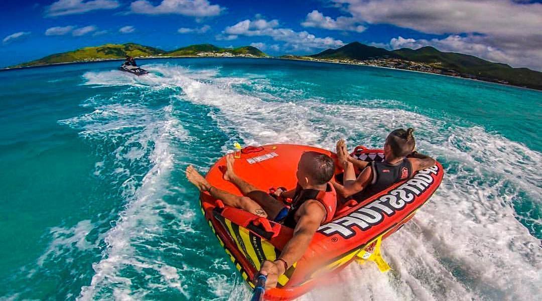 Two people on an inflatable tube being pulled across turquoise water by a boat.
