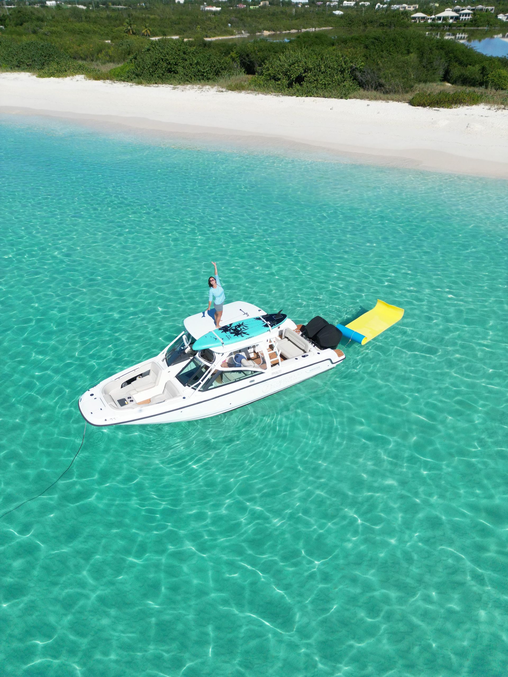A boat on clear turquoise water near a white sand beach. A person stands on the boat; a yellow float trails behind.