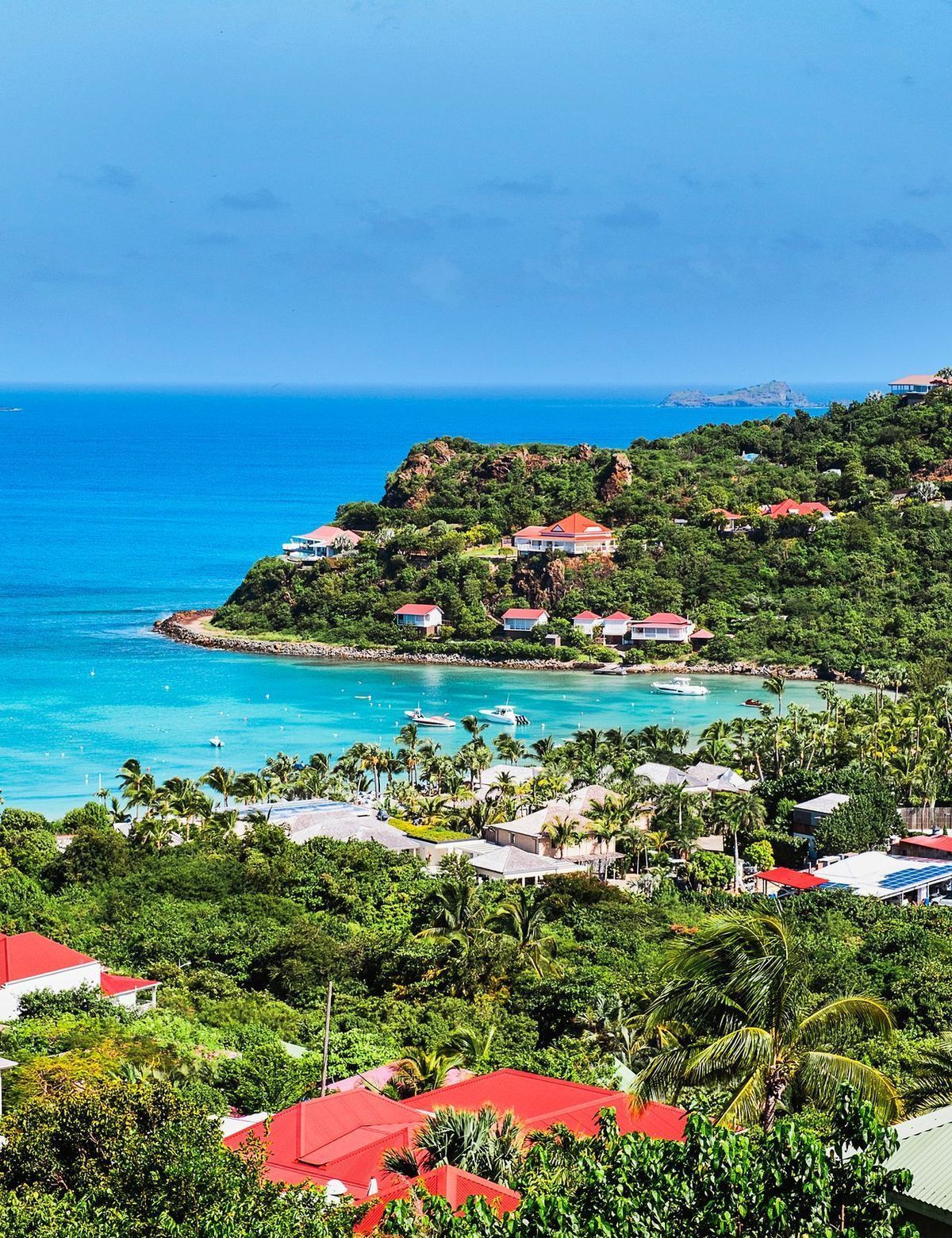 Tropical coastal view with turquoise water, lush green hillside, and villas with red roofs.