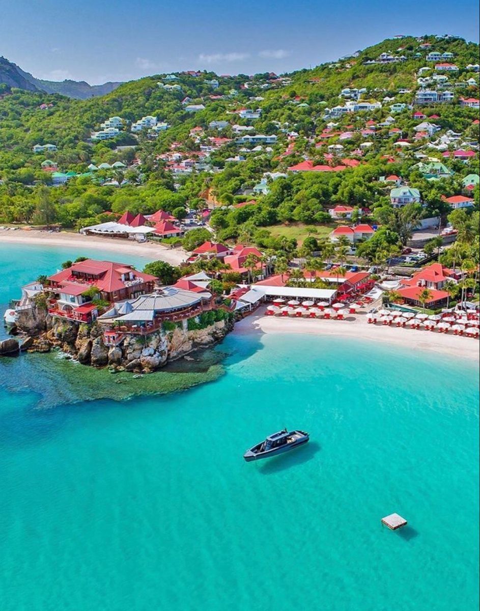 Tropical beach scene with turquoise water, buildings on a hillside, and red-roofed structures along the shore.