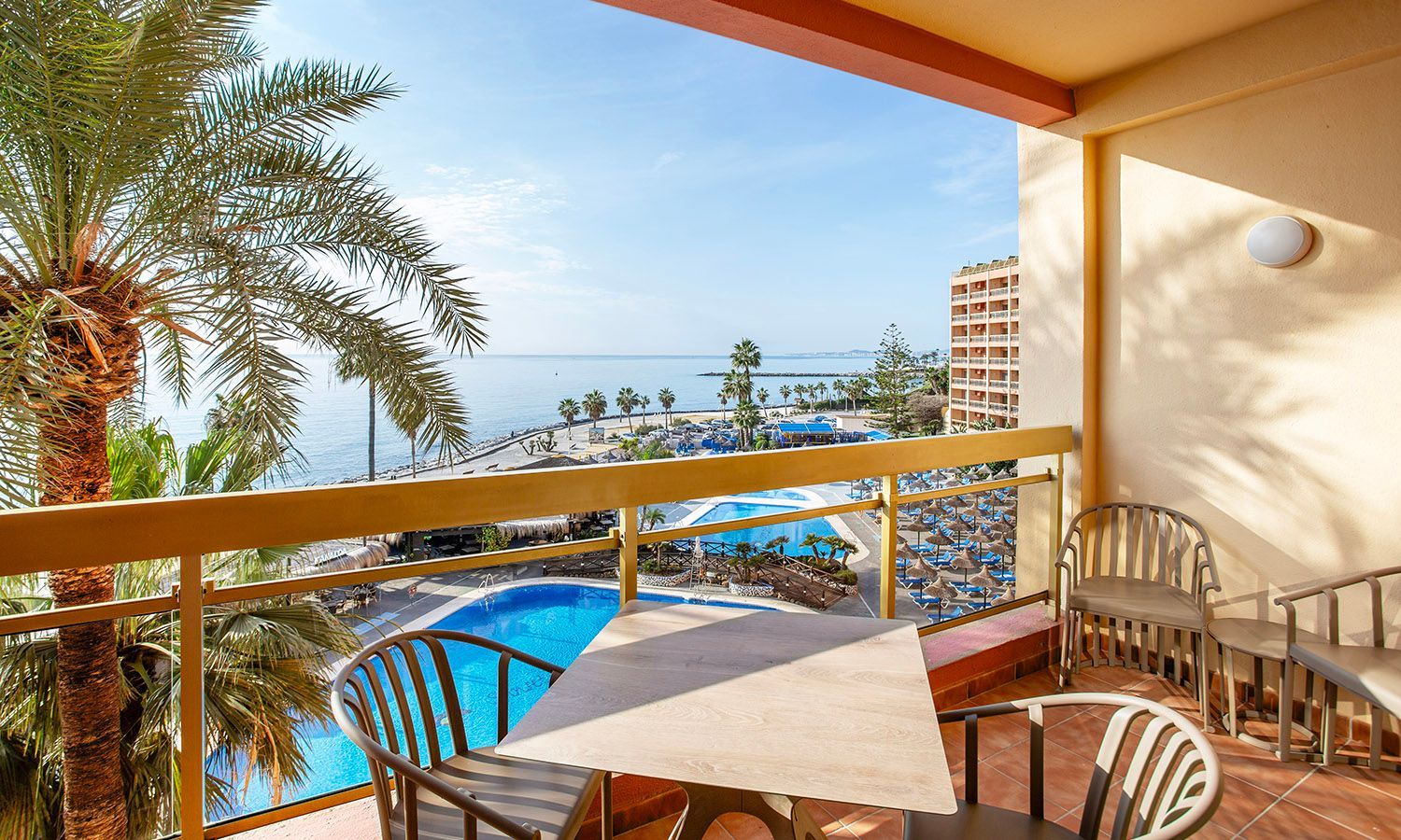 Balcony overlooking a resort pool and ocean, with palm trees and a table and chairs in the foreground