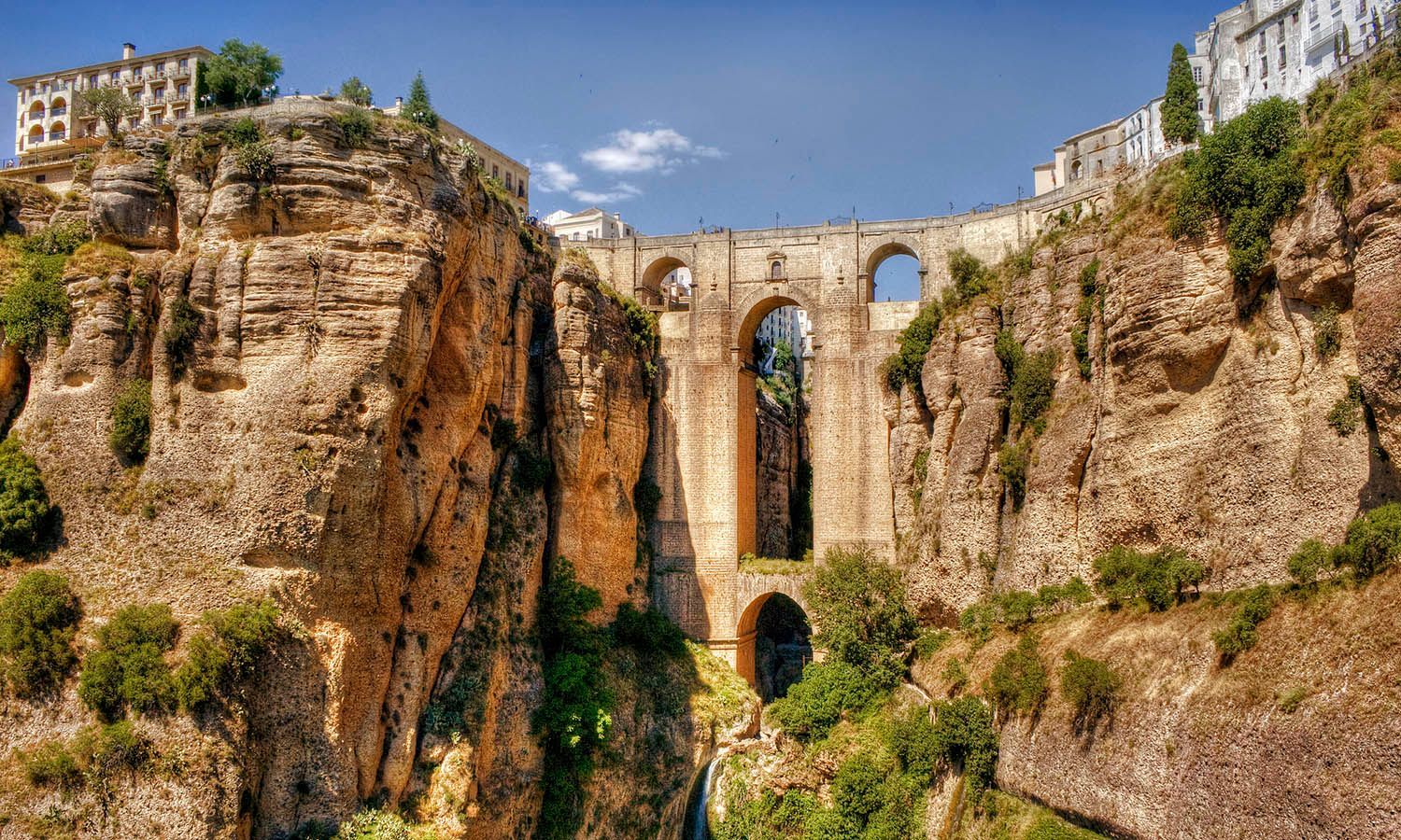 Historic stone bridge spanning a deep rocky gorge beside cliffside buildings under a blue sky