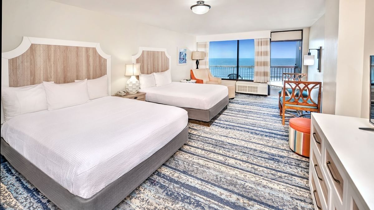 Bright hotel room with two beds, blue patterned carpet, and ocean-view windows