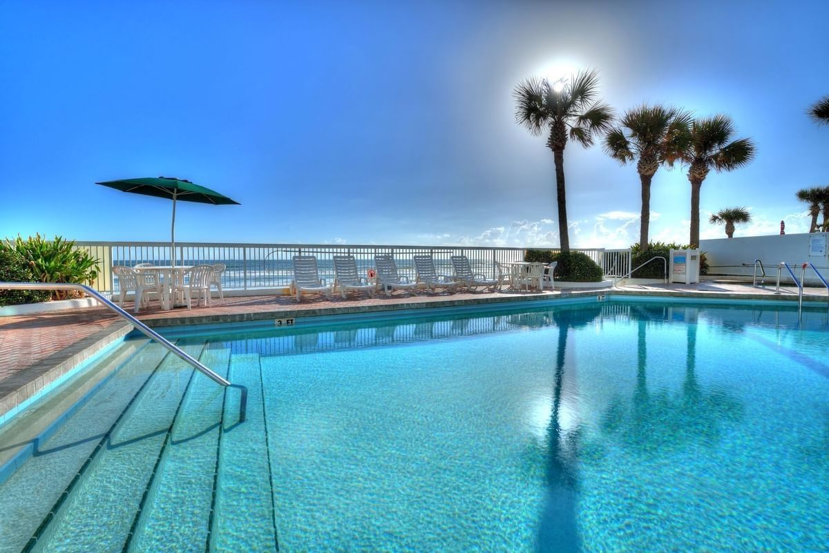 Poolside scene with clear pool, palm trees, and ocean view under bright sun.