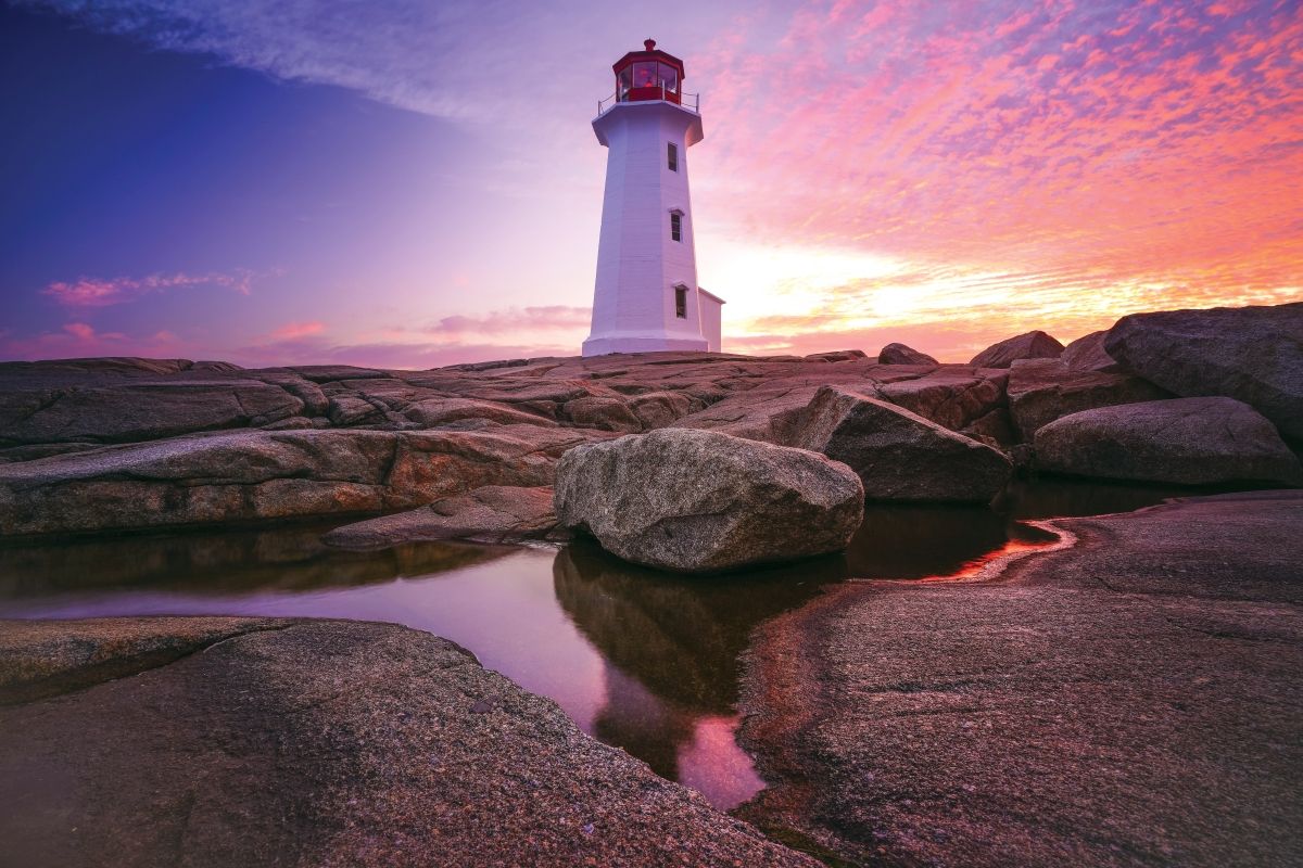 Lighthouse at sunset, white tower with red top, rocky shoreline, sky with pink and purple hues.