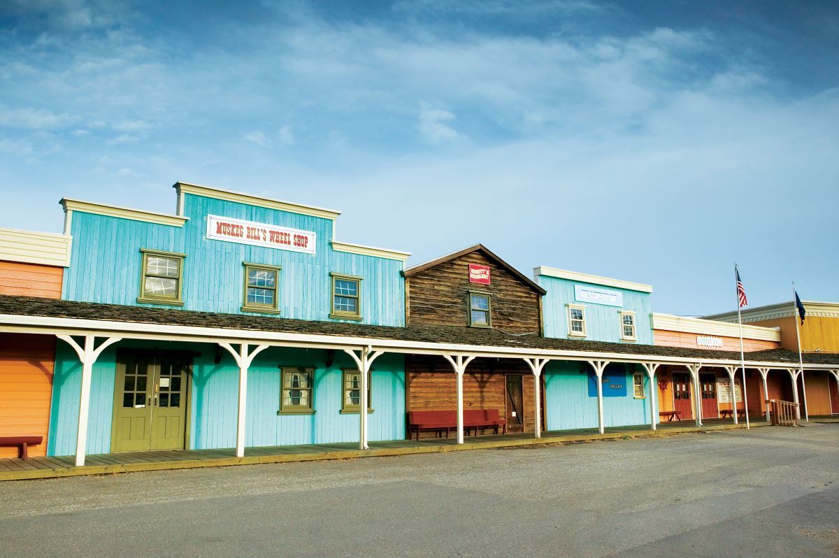 Western town storefronts with turquoise, brown, and orange buildings under a blue sky.