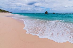 Pink sand beach, turquoise water, white waves. Cloudy sky, distant rock formations.