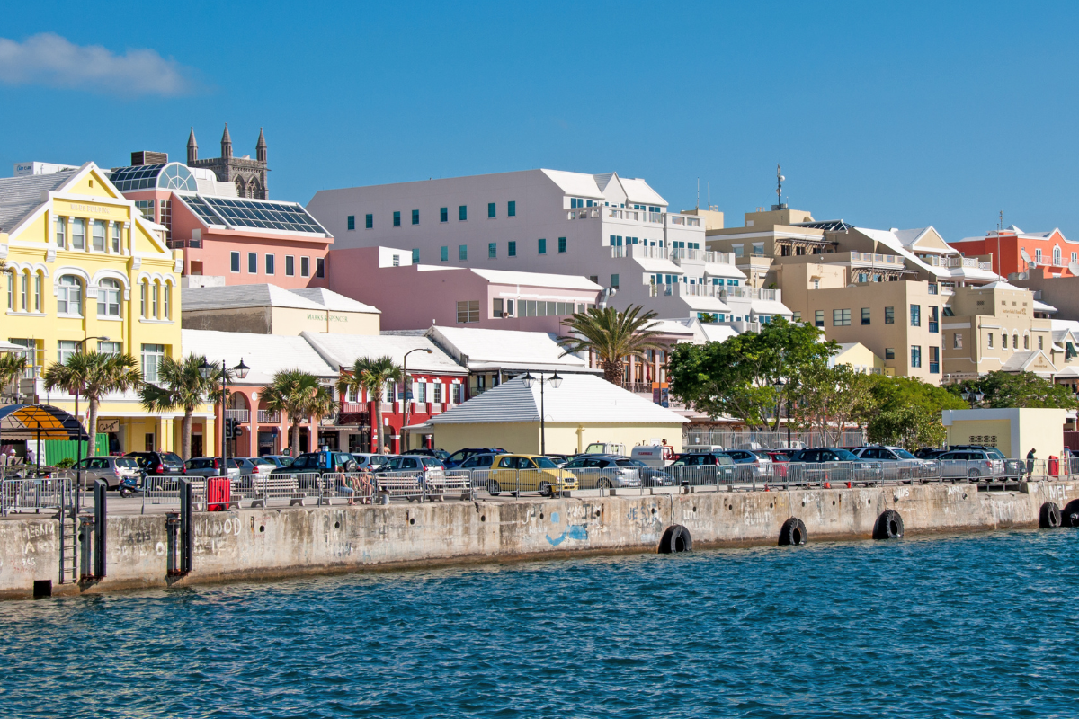Colorful colonial-style buildings line a waterfront in Hamilton, Bermuda, with a row of parked cars along the concrete quay.