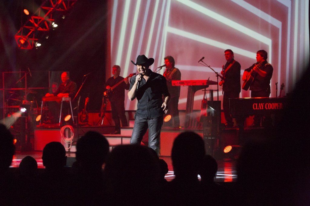 A country singer in a cowboy hat performs on stage with a band, red stage lighting, and a crowd.