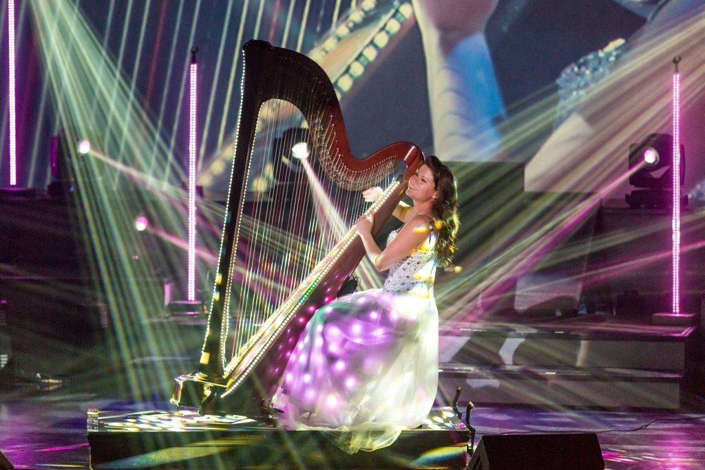 Woman in sparkling gown playing a harp on a stage with spotlights and a screen in the background.