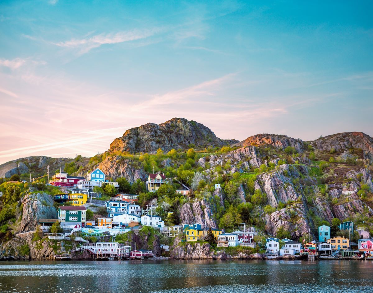 Colorful houses nestled against a rocky hill by the water under a soft, blue sky.