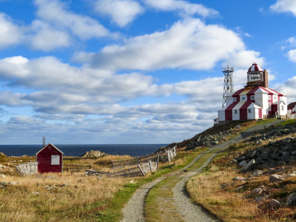 Red and white lighthouse on a hill overlooking the ocean. A path leads to the building, and a red shed sits below.