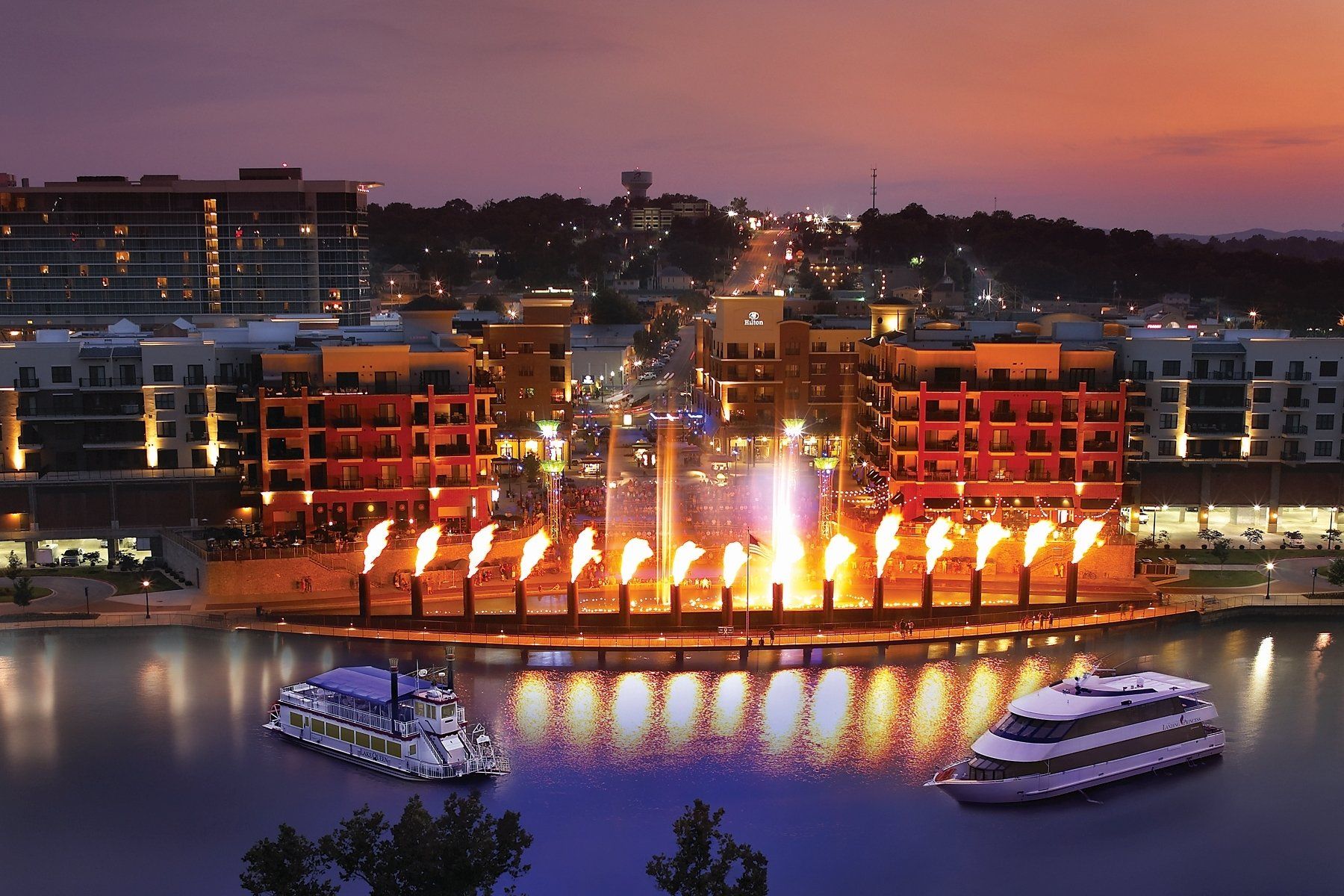 Fountain with flames and city buildings at dusk, reflected in water with two boat cruises.