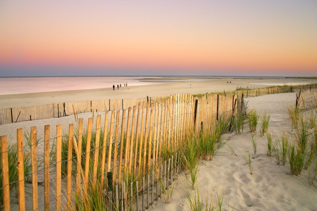 Beach at dusk with a wooden fence, sandy dunes, and colorful sky.