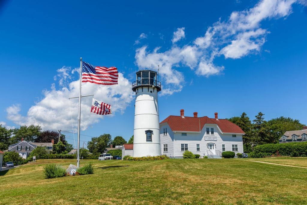 Lighthouse with American flags on a grassy hill under a blue sky, with a white building and trees in the background.