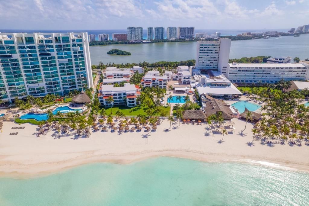 Aerial view of a beach resort in Cancun with white sand, turquoise water, palm trees, and tall buildings.