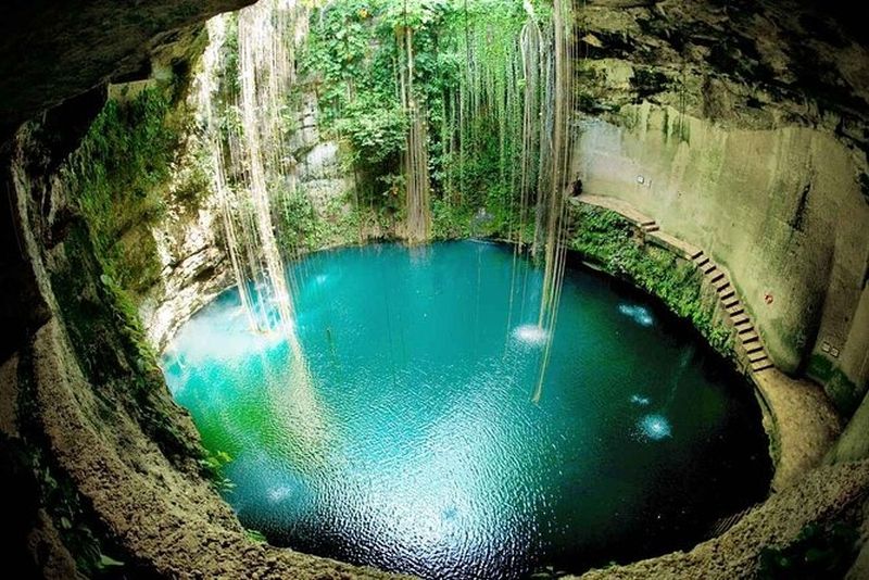 Circular cenote with turquoise water, surrounded by limestone walls and greenery; light streams down.