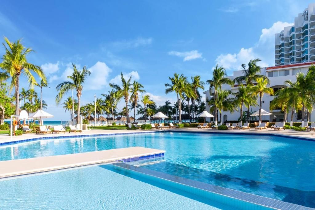Large pool at a resort with palm trees, sunny skies, and a view of the ocean.