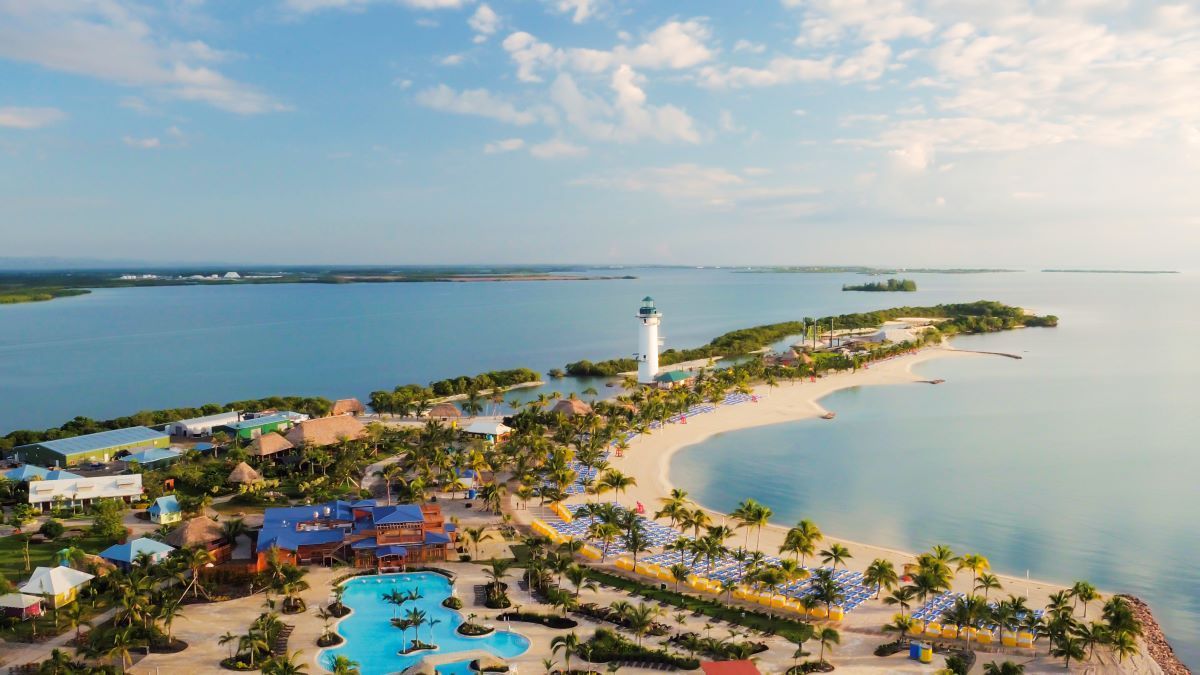 Aerial view of an island with a lighthouse, beach, and resort buildings; turquoise water and blue sky.