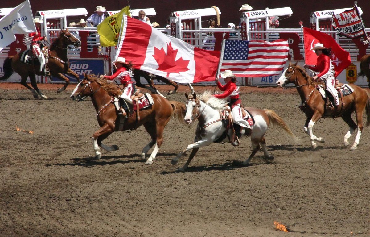 Cowboys on horses carry flags of Canada, the US, and others at a rodeo.