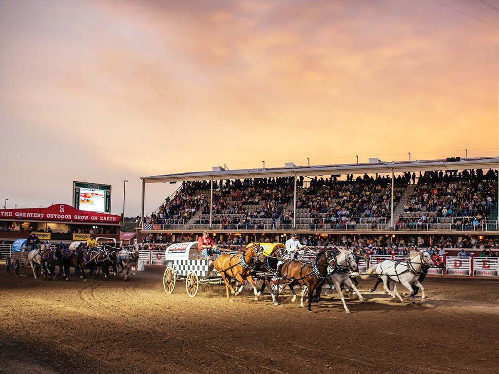 Sunset rodeo scene with horse-drawn wagons racing around a dirt track, crowd in the stands.