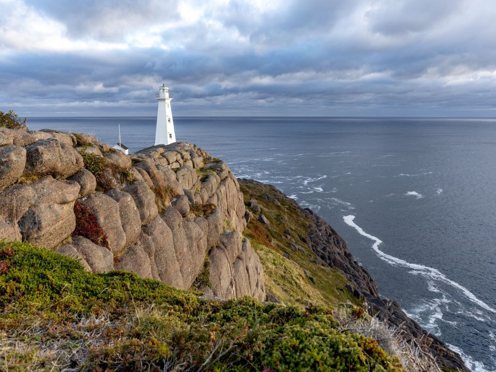 White lighthouse on rocky cliff overlooking the ocean under a cloudy sky.