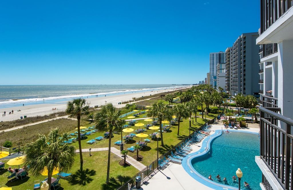 Oceanfront view of a beach, pool, and buildings with palm trees under a bright blue sky.