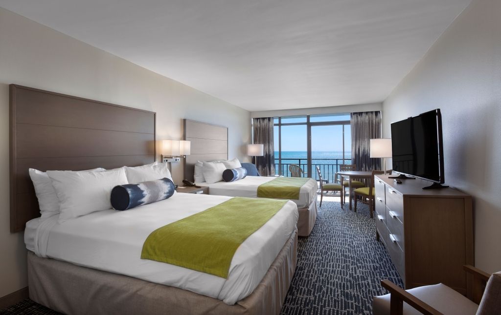 Hotel room with two double beds, ocean view through sliding doors, TV, neutral tones.