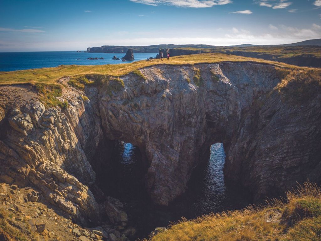 Coastal cliffs with two large sea caves, blue water, and grassy terrain.