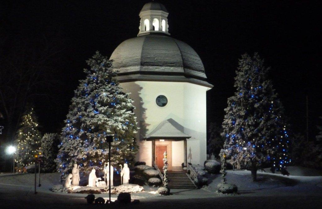 A snow-covered white chapel at night, decorated with Christmas lights and greenery.