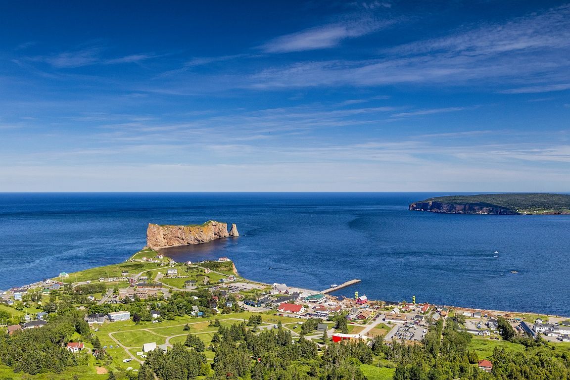 Coastal view: Village nestled along water, large rock formation in sea, blue sky.