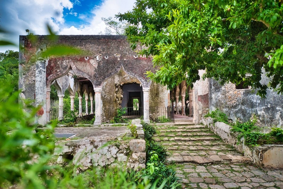 Ruined stone building with arched openings, steps, and overgrown foliage; jungle setting.
