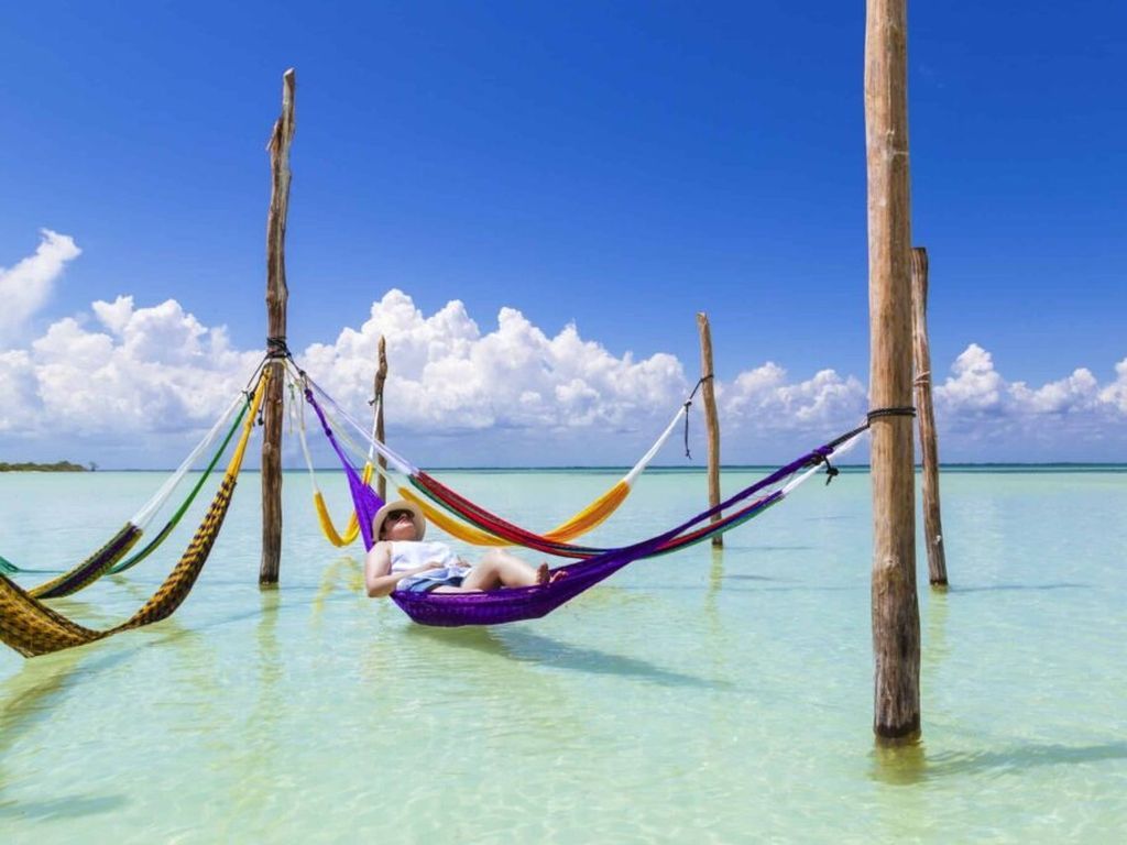 Person relaxing in a purple hammock in shallow turquoise water under a blue sky.