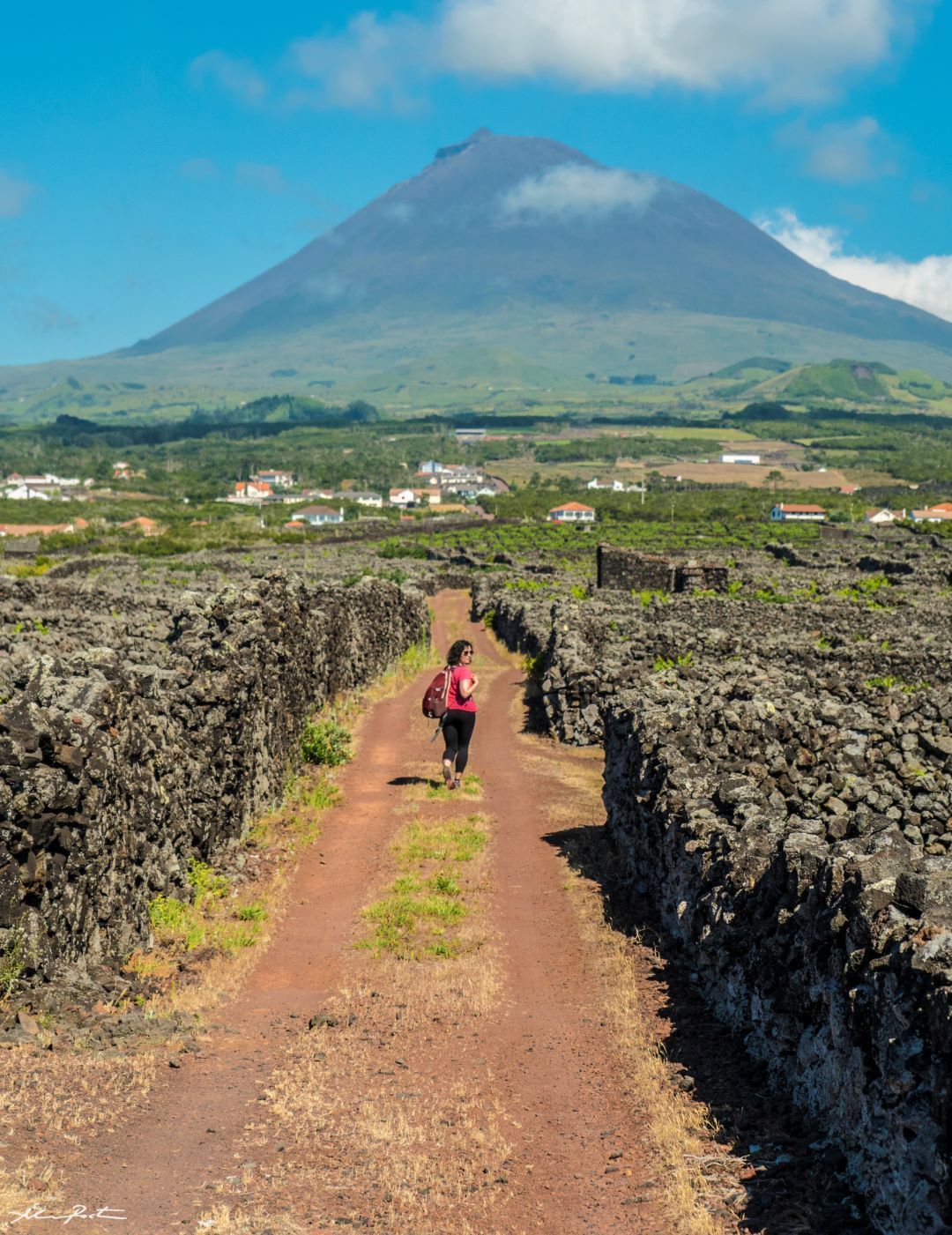 Person walking a dirt path between lava walls toward a green mountain under a blue sky