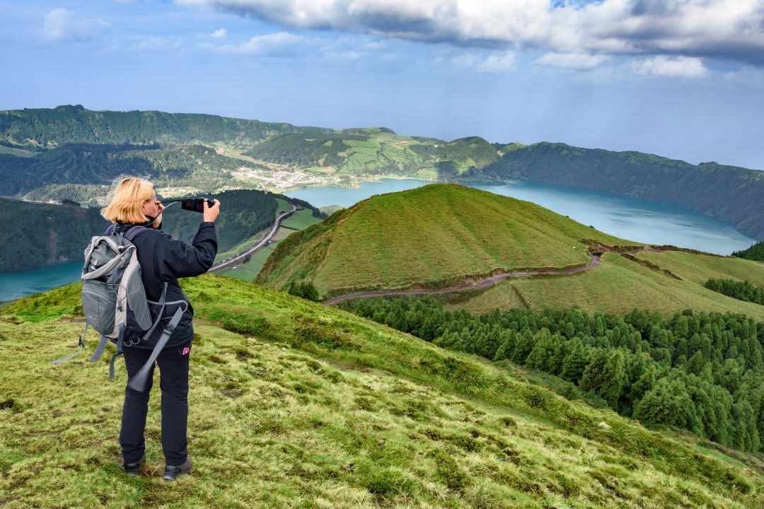 Hiker with backpack photographing a green volcanic ridge and blue bay under cloudy skies