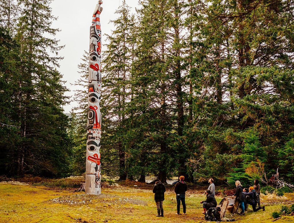 Tall painted totem pole in a forest clearing with several people standing and sitting nearby