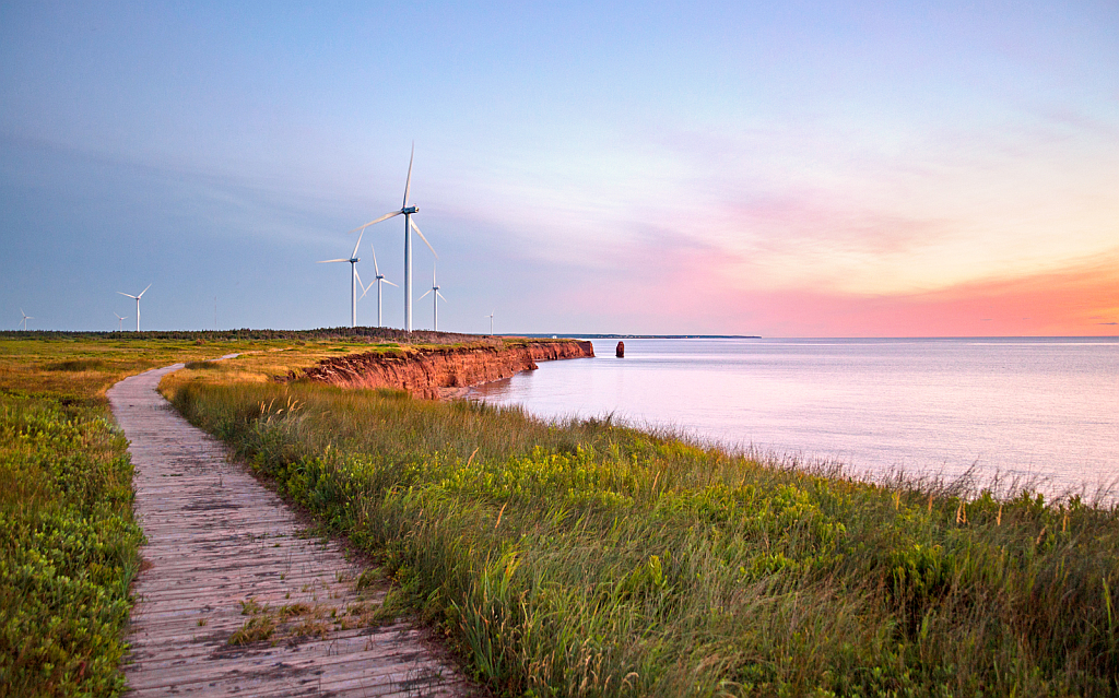 Path beside a body of water, wind turbines in the distance at sunset with pink and orange sky.