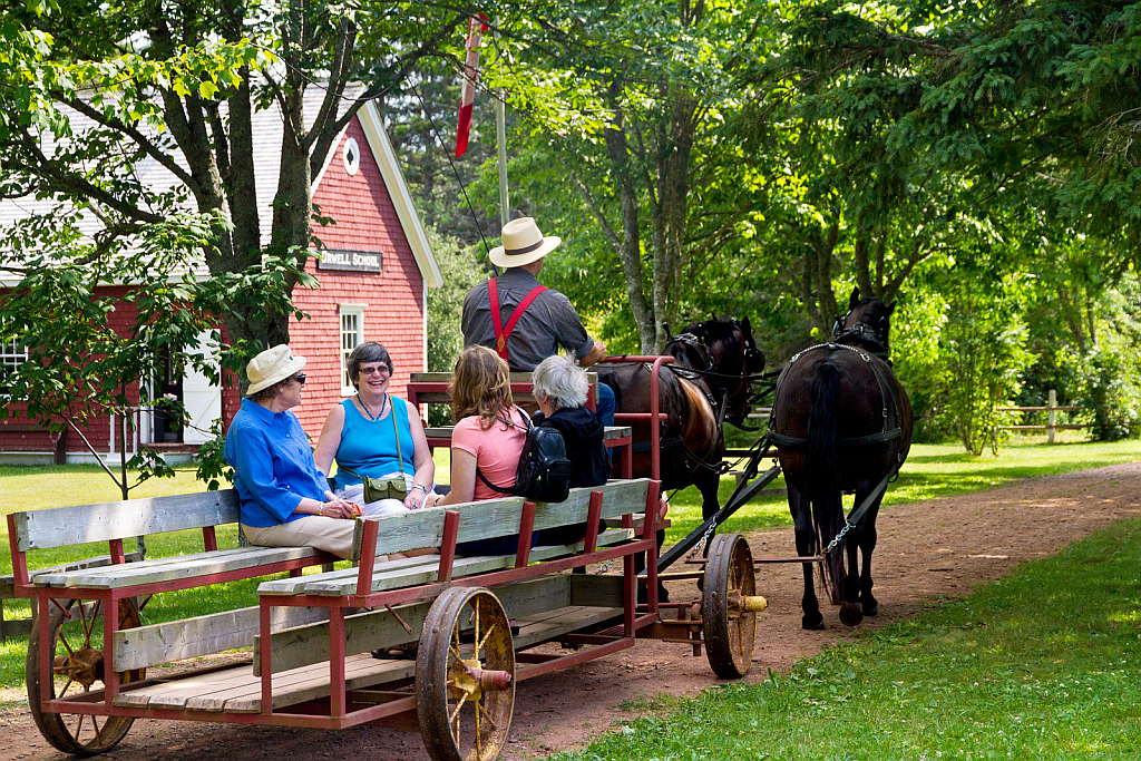 Horse-drawn wagon with passengers on a dirt path beside a red building and lush greenery.