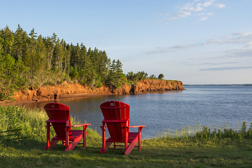 Two red Adirondack chairs face a calm body of water, near a red cliff and lush green trees.