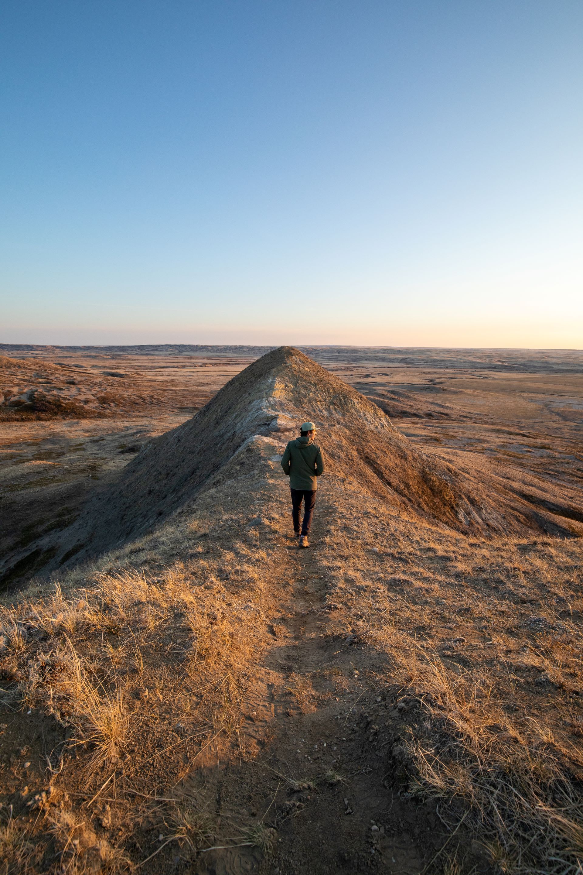 Person walking along a dirt ridge in a vast dry landscape under a clear blue sky