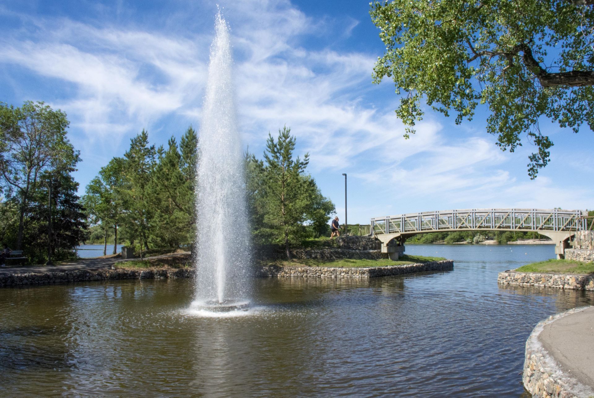 Fountain spraying high into the air in a lake, with a bridge and trees in the background under a blue sky.