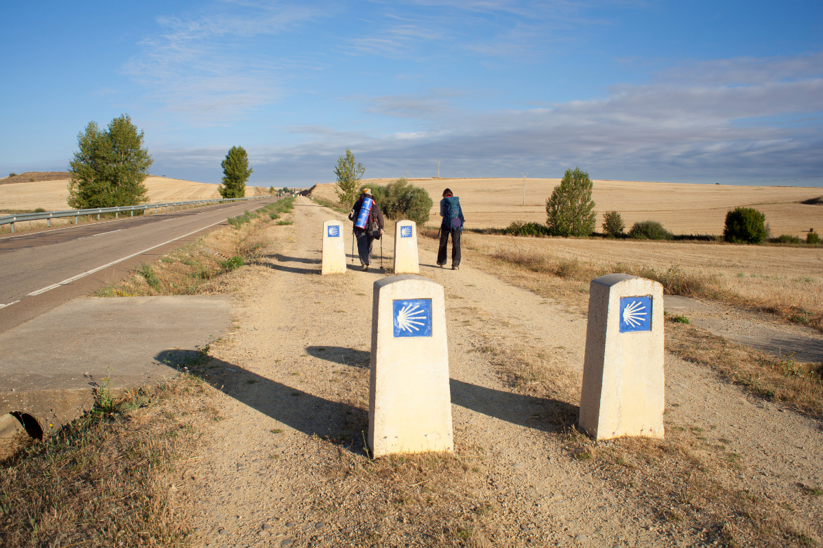 Two people walking on a rural path near roadside signs and beige posts under a blue sky
